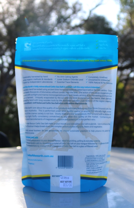 "Back view of a blue and white resealable pouch of Celtic Sea Salt, placed outdoors. The label describes the product as fully mineralised and naturally harvested sea salt, free from anti-caking agents and additives. It highlights its natural source, traditional harvesting methods, and health benefits. The packaging also includes usage tips, nutritional claims, a barcode, and a best-before date of December 2030."