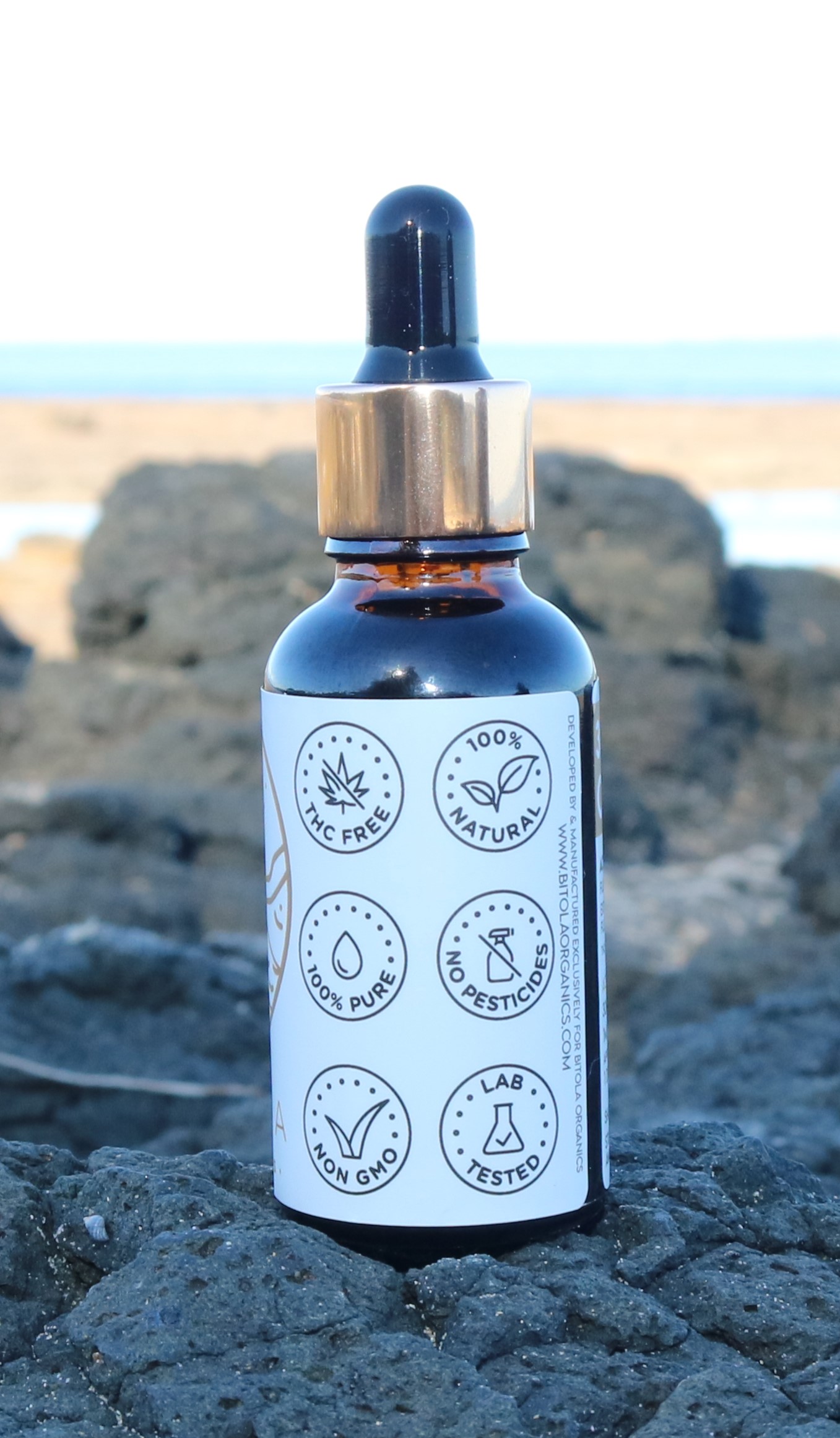 A dark amber dropper bottle with a gold cap, labeled with six icons indicating key product features: THC Free, 100% Natural, 100% Pure, No Pesticides, Non-GMO, and Lab Tested. The bottle is placed on black volcanic rock near the beach, with a blurred background of sand, sea, and sky.