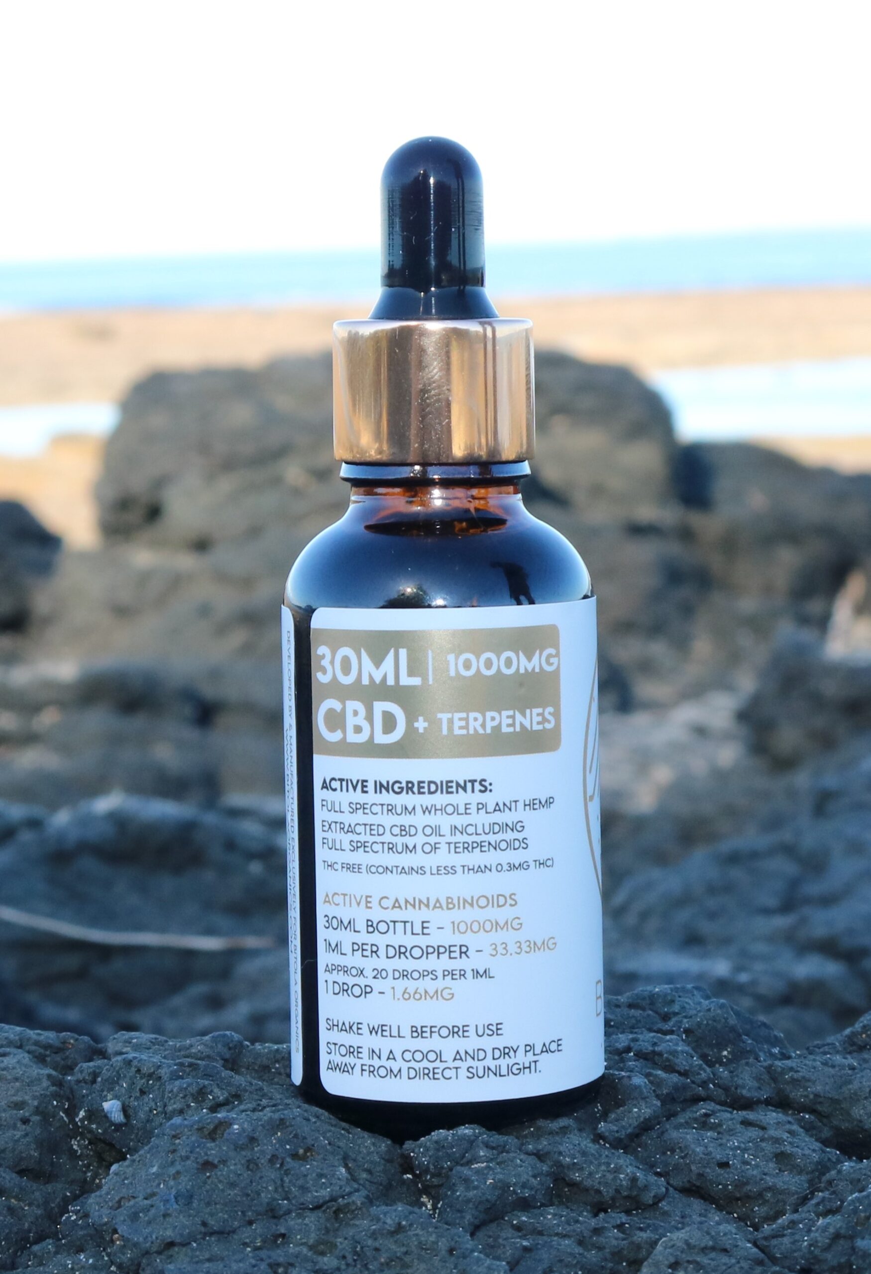 A 30ml amber glass dropper bottle labeled "CBD + Terpenes 1000mg." The label details include active ingredients such as full-spectrum whole plant hemp extracted CBD oil and terpenoids. It states the product is THC-free (containing less than 0.3% THC). Dosage information indicates 33.33mg per 1ml dropper and 1.66mg per drop. The bottle is placed on black volcanic rock with a blurred background of a coastal scene.