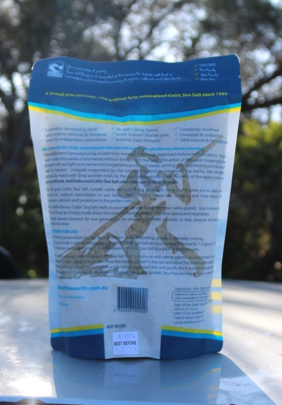 Back view of a packet of Celtic Sea Salt placed outdoors on a flat surface, with trees and sunlight in the background. The packaging contains product information, including benefits such as being hand-harvested, unrefined, free of anti-caking agents, and rich in trace minerals. A barcode and “Best Before” label are visible near the bottom.