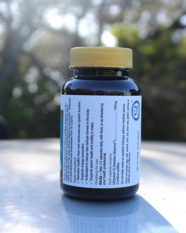 "A dark-colored supplement bottle with a yellow cap placed outdoors on a reflective surface. The label on the bottle includes dosage instructions and supplement facts, including 'Ubidecarenone (Co-Enzyme Q10 – Ubiquinol)' with a dosage of 150mg. The background is softly blurred with trees and sunlight visible."