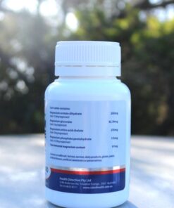A white plastic bottle of magnesium supplement photographed outdoors on a reflective surface, with trees and sunlight in the background. The label lists magnesium compounds and their respective quantities, and includes the company name 'Health Direction Pty Ltd' along with contact details and website.