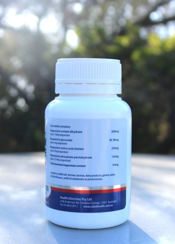 A white plastic bottle of magnesium supplement photographed outdoors on a reflective surface, with trees and sunlight in the background. The label lists magnesium compounds and their respective quantities, and includes the company name 'Health Direction Pty Ltd' along with contact details and website.